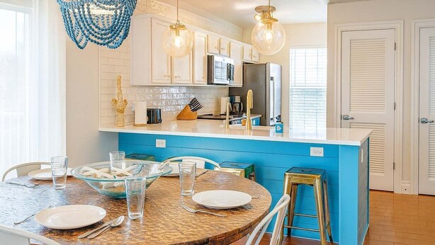 A modern kitchen with white cabinets, a blue island, and a dining area with a round table and chairs. - Sun Dancer #11 2 Bedroom Home by RedAwning (South Padre Island)