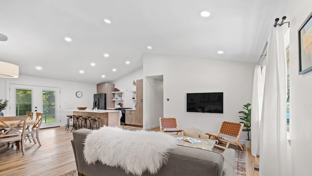 Un salon moderne avec un canapé gris, un plaid blanc et moelleux et une table basse. On aperçoit une cuisine en arrière-plan, avec des meubles en bois et un téléviseur fixé au mur.