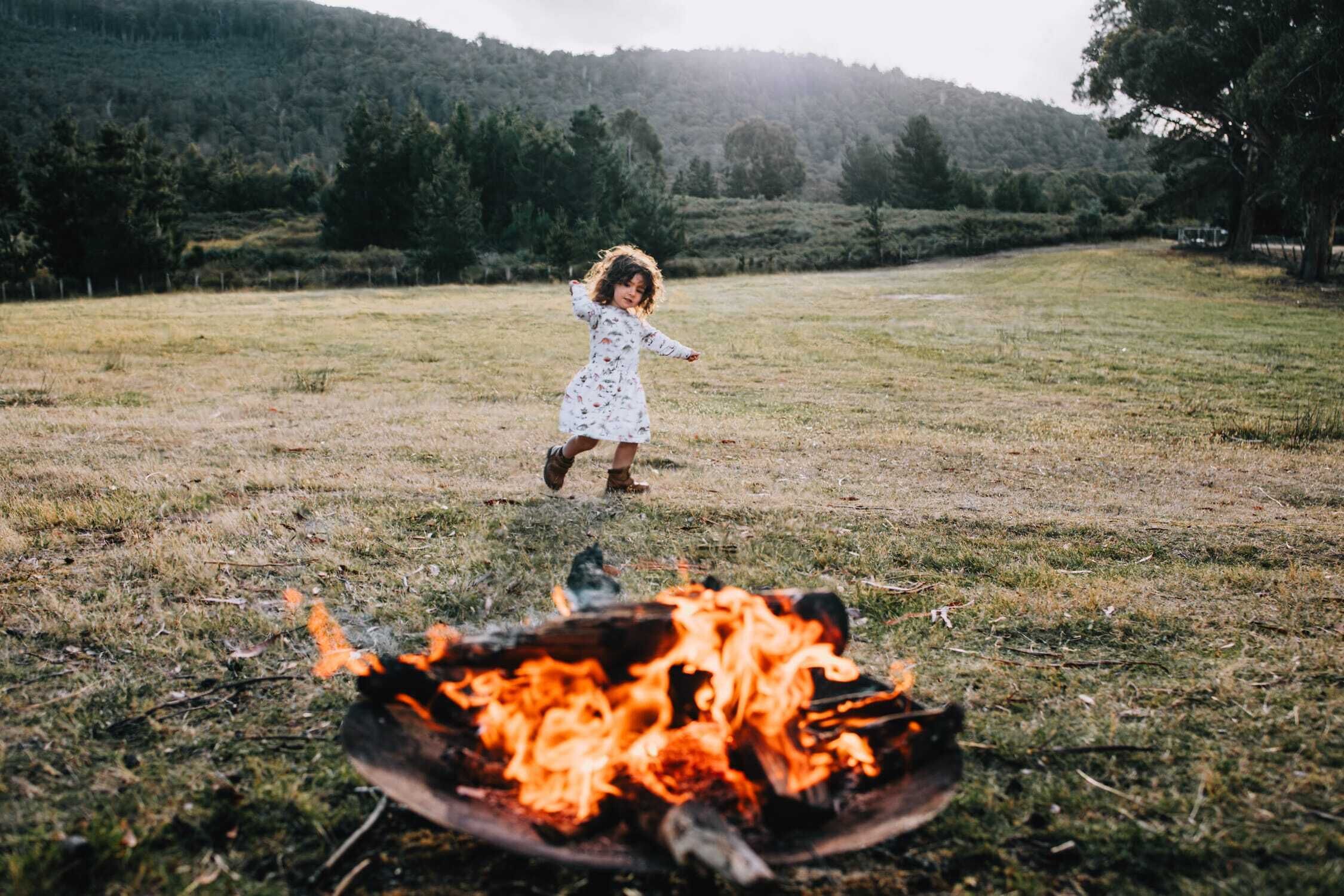 A child playing near a campfire in a grassy field with trees in the background.