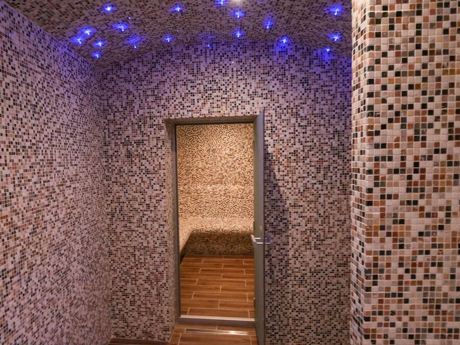 A narrow hallway with mosaic walls and a wooden floor, leading to a room with a tiled floor.