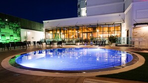 A swimming pool with a glass enclosure, surrounded by outdoor seating and a building with large windows.