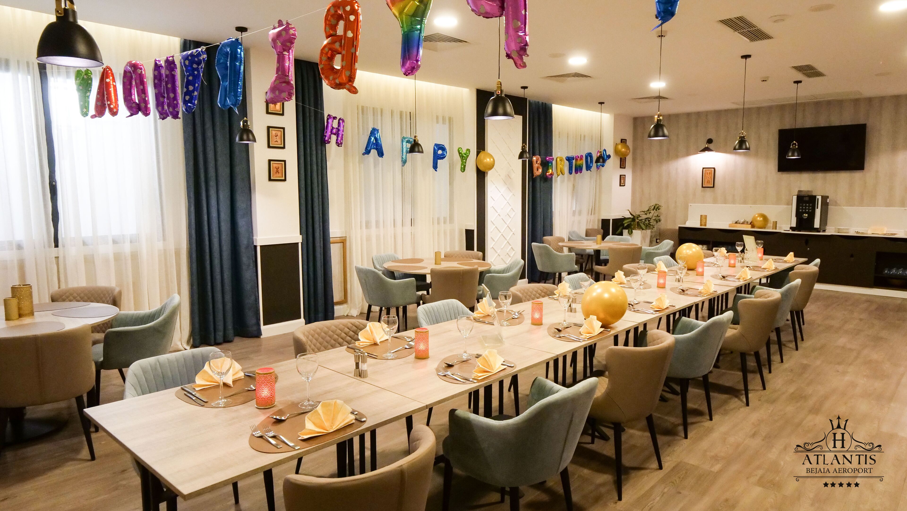 Un área de comedor con varias mesas dispuestas para una celebración, globos que deletrean “HAPPY BIRTHDAY”, y un letrero que indica “Atlantis Bejaia Airport”.