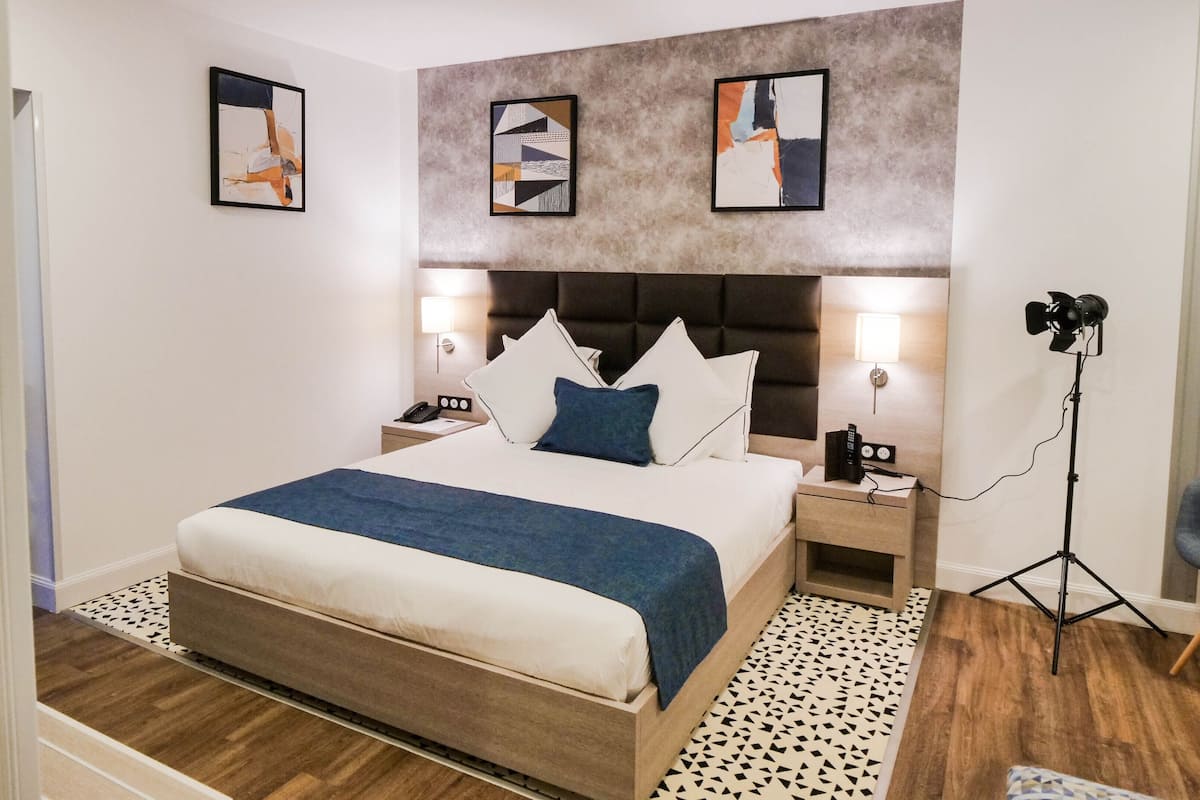 A modern hotel room with a large bed, two bedside tables, a desk, and a camera on a tripod.