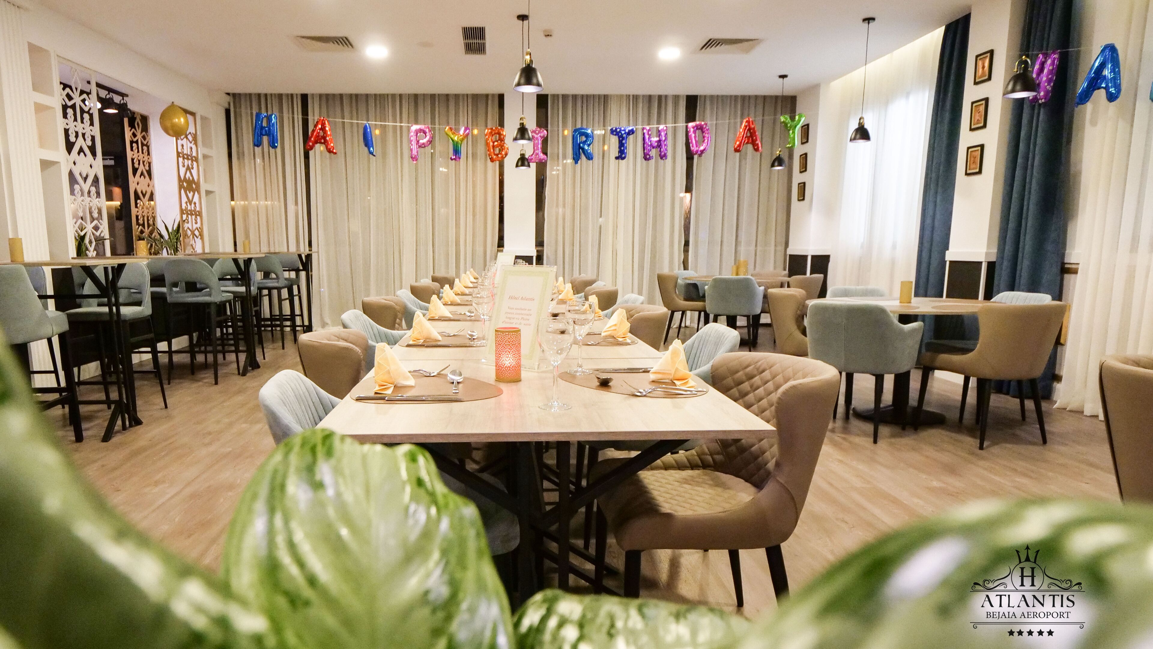 A banquet hall with tables set for a birthday celebration, featuring a banner with balloons spelling out "HAPPY BIRTHDAY".
