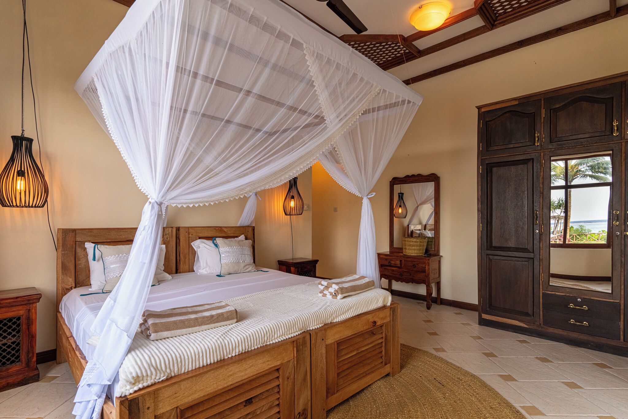 A bedroom with a wooden bed, white bedding, a canopy, bedside lamps, a wooden wardrobe, and a view through a window.
