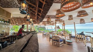 A beachside restaurant with wooden furniture, wicker hanging lamps, and a view of the sea. - Marafiki Beach Hotel & Spa (Matemwe)
