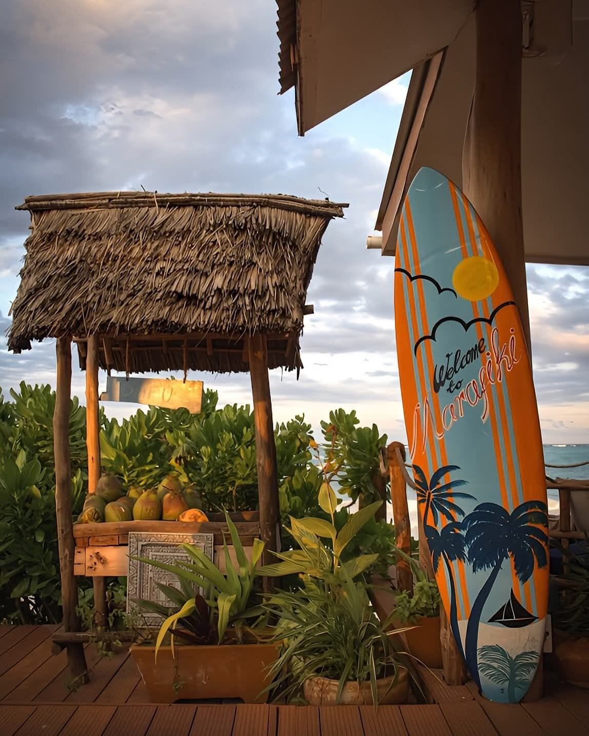 A wooden structure with a thatched roof, a surfboard with a tropical design, and potted plants.
