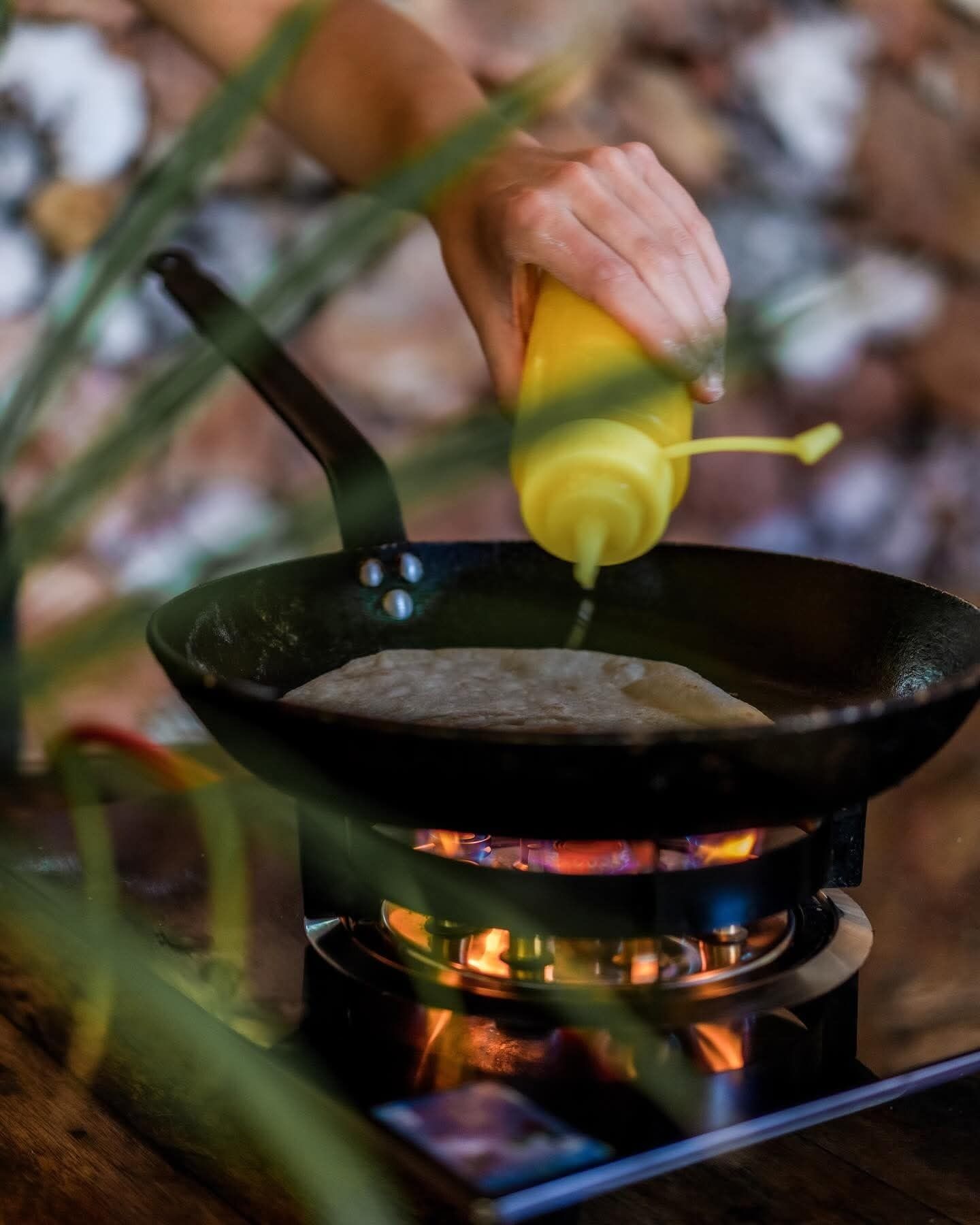 A hand pouring a yellow liquid into a black frying pan on a portable stove.
