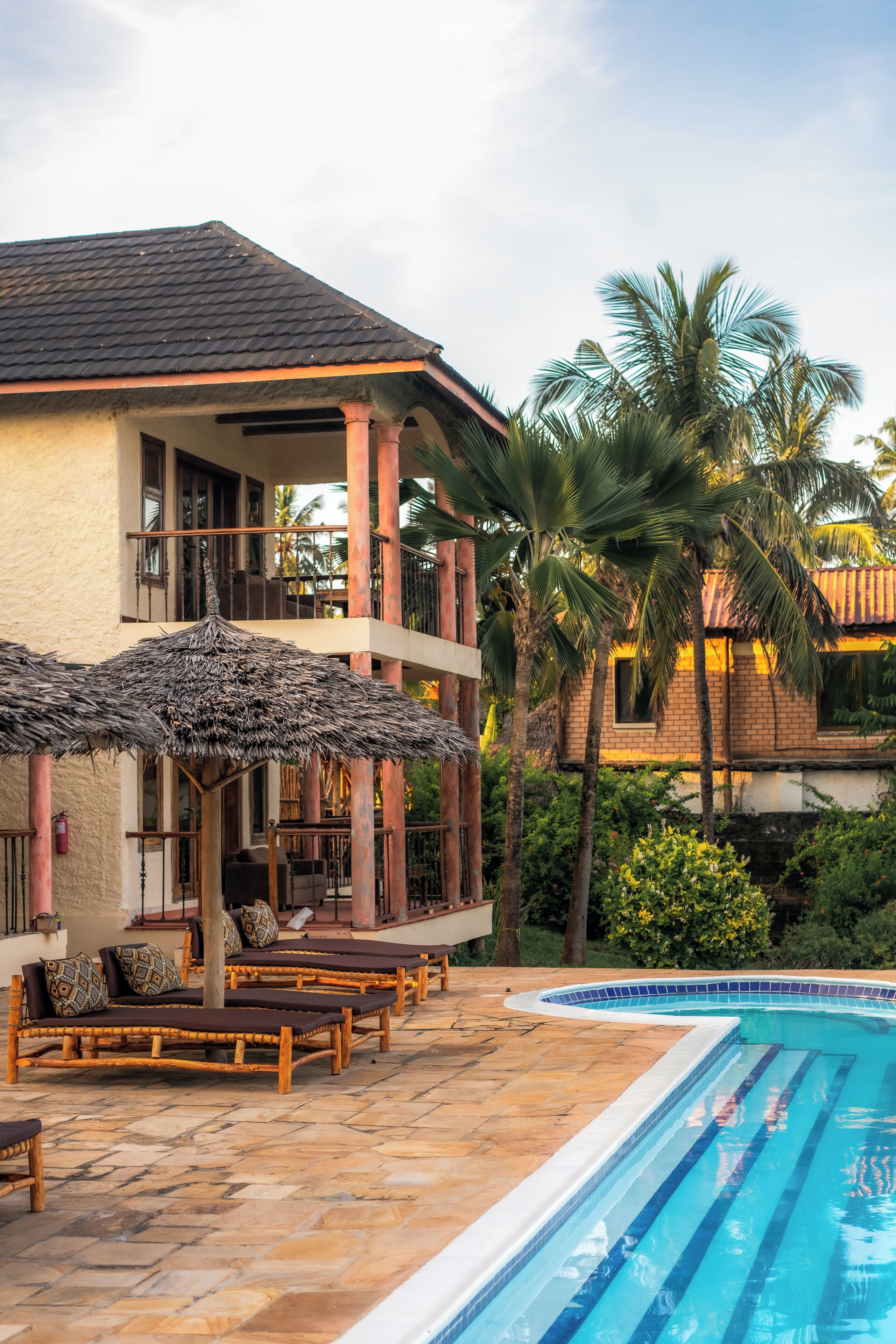 A pool area with lounge chairs and a thatched umbrella, surrounded by palm trees and a building with a tiled roof.
