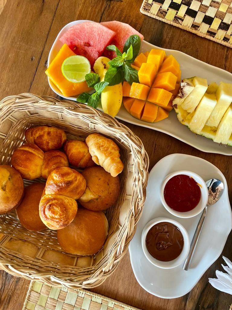 A table with a basket of bread rolls, a plate of sliced fruits, and two small bowls of sauce.