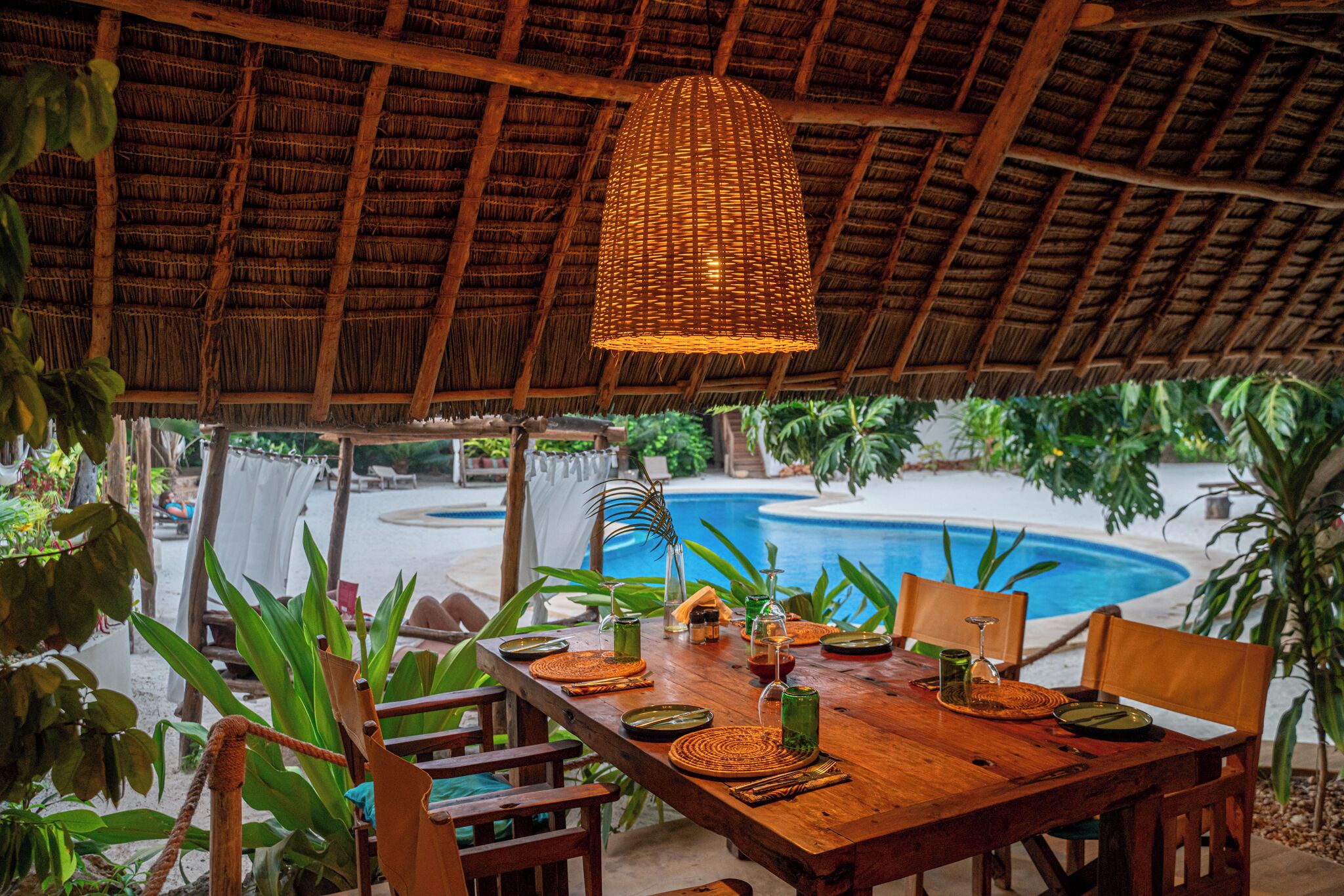 A wooden dining table set with plates, glasses, and a hanging light fixture in a tropical setting with a pool in the background.