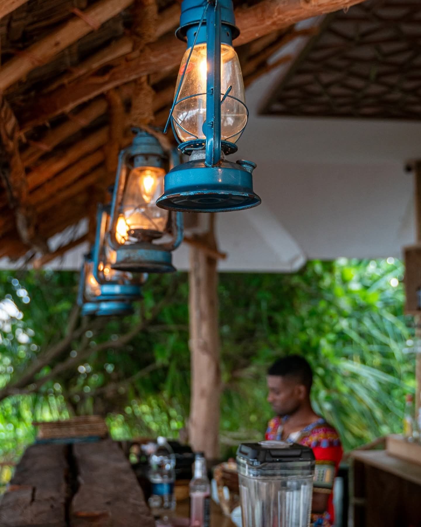 Un espacio de comedor al aire libre con estilo rústico, faroles colgantes y una persona al fondo.