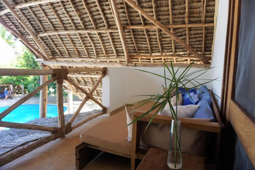 A room with a thatched roof, a wooden bench, a glass vase with a plant, and a view of a swimming pool.