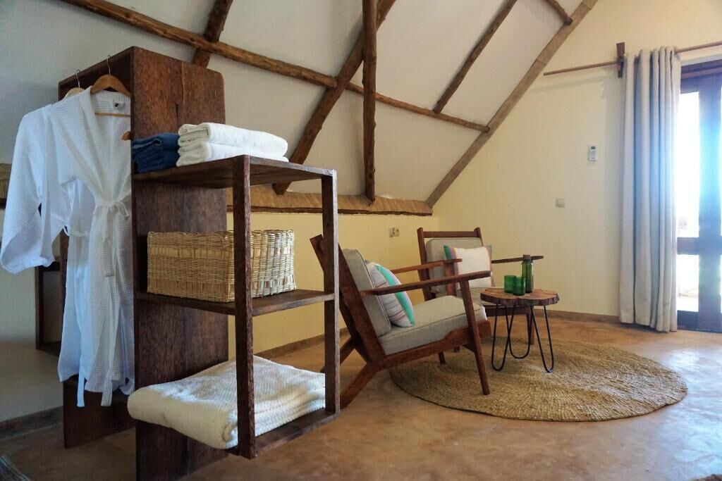 A rustic room with a wooden shelf holding folded clothes, a wicker basket, and a chair with cushions. There's a small round table with a green candle.