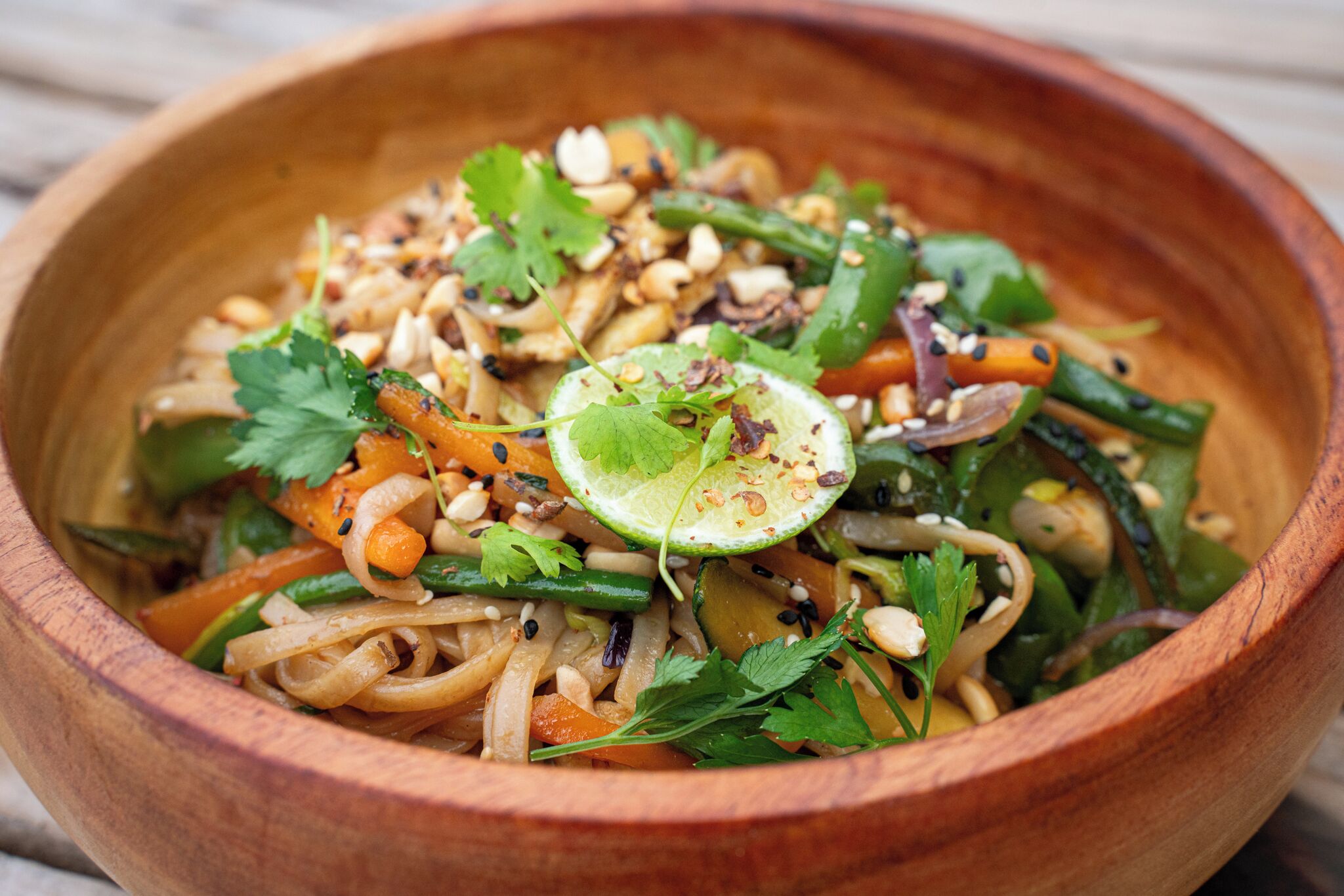 A wooden bowl containing a noodle dish with vegetables and herbs.