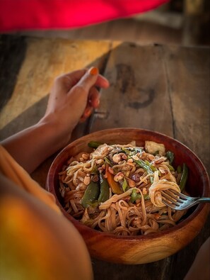 A bowl of noodles with vegetables and nuts, held by a person with orange nails. - Marafiki Beach Hotel & Spa (Matemwe)