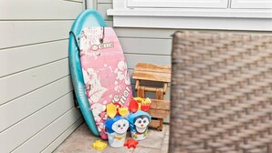Une planche de surf appuyée contre un mur, un petit tabouret en bois et une chaise en osier.