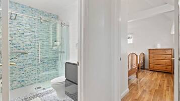 A bathroom with a glass shower enclosure, a toilet, and a wooden dresser.