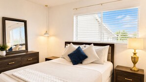 Un dormitorio ordenado con cama, mesitas de noche, lámpara, espejo y ventana con persianas.