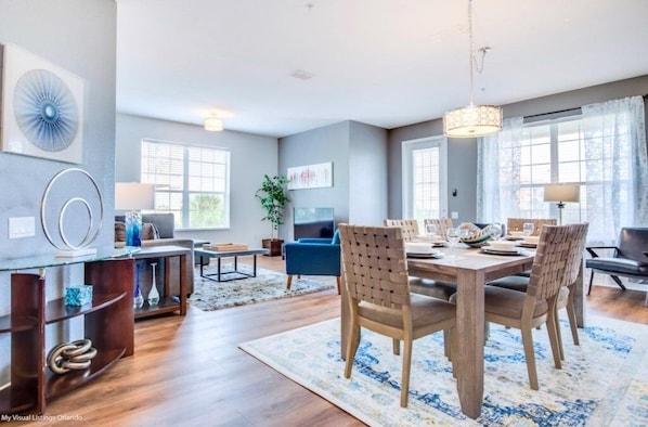 A modern dining area with a wooden table and chairs, a chandelier, and a large window with curtains. - Vista Cay Electra 3 Bedroom Condo by RedAwning (Orlando)