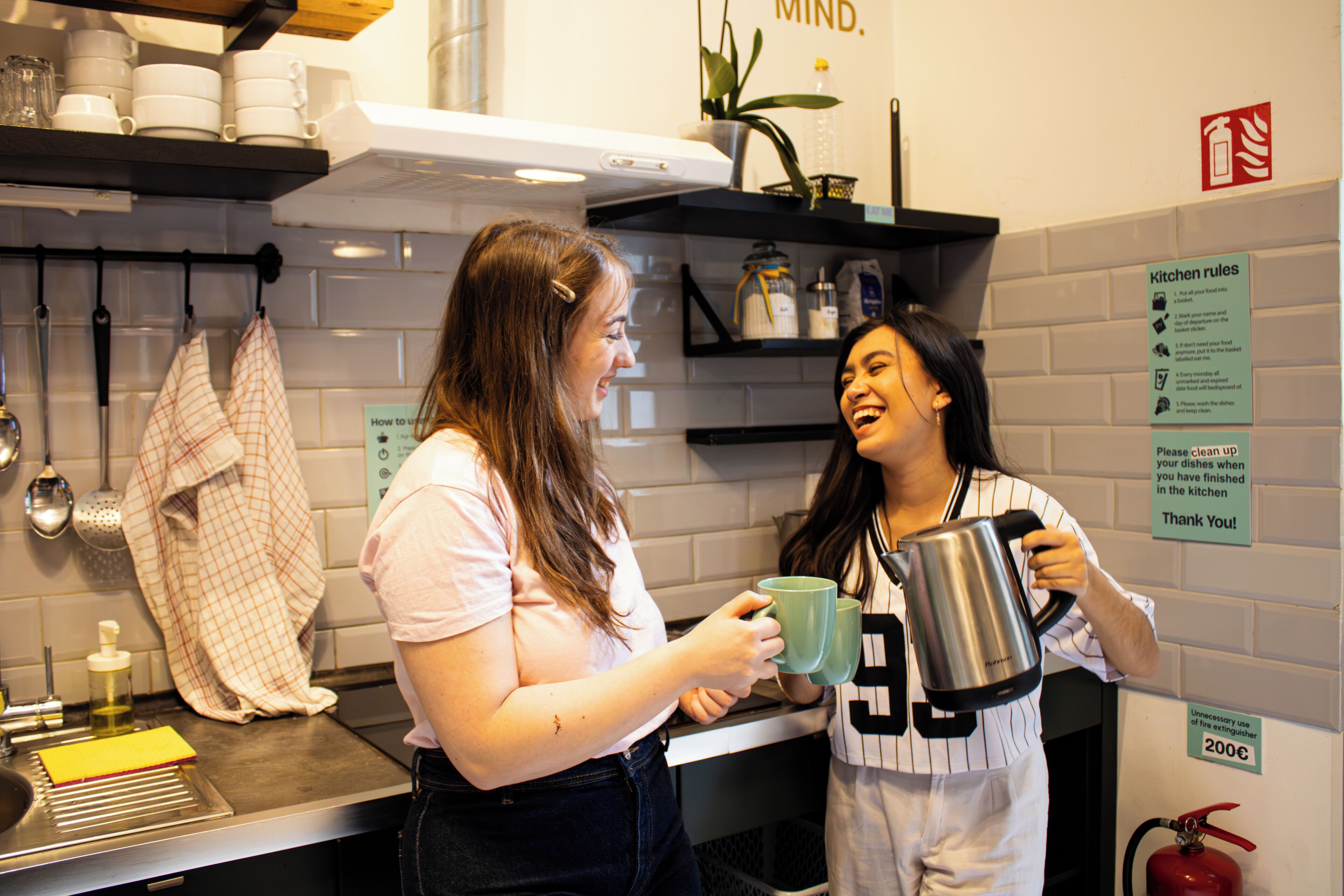 Deux femmes dans une cuisine, tenant des tasses et une bouilloire.
