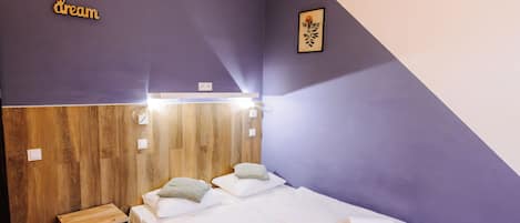 Un dormitorio con suelo de madera, una cama con sábanas blancas, una mesita de noche de madera y una lámpara fijada a la pared.