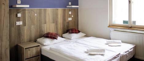 Un dormitorio con cabecera de madera, dos camas, una mesita de noche, un ventanal con vistas y un radiador.