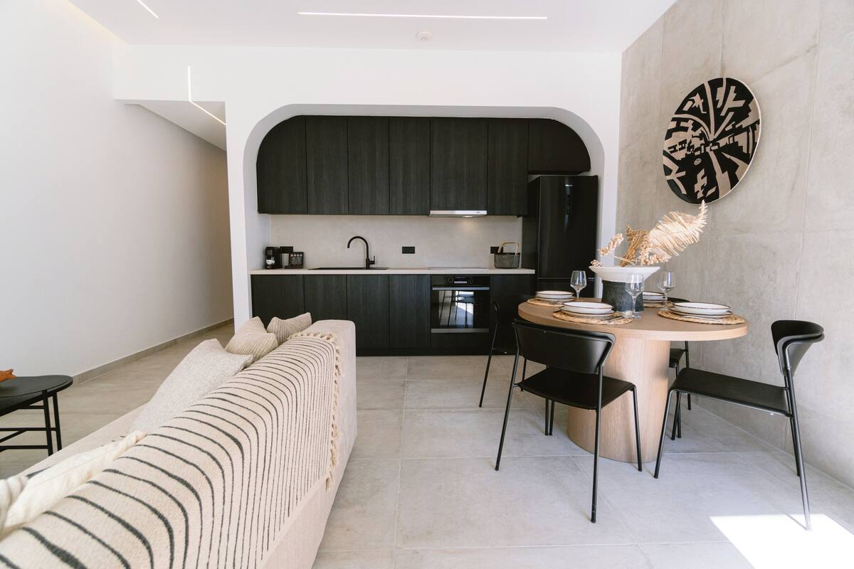A modern kitchen with dark cabinets, a built-in oven, and a dining area with a round table and chairs.