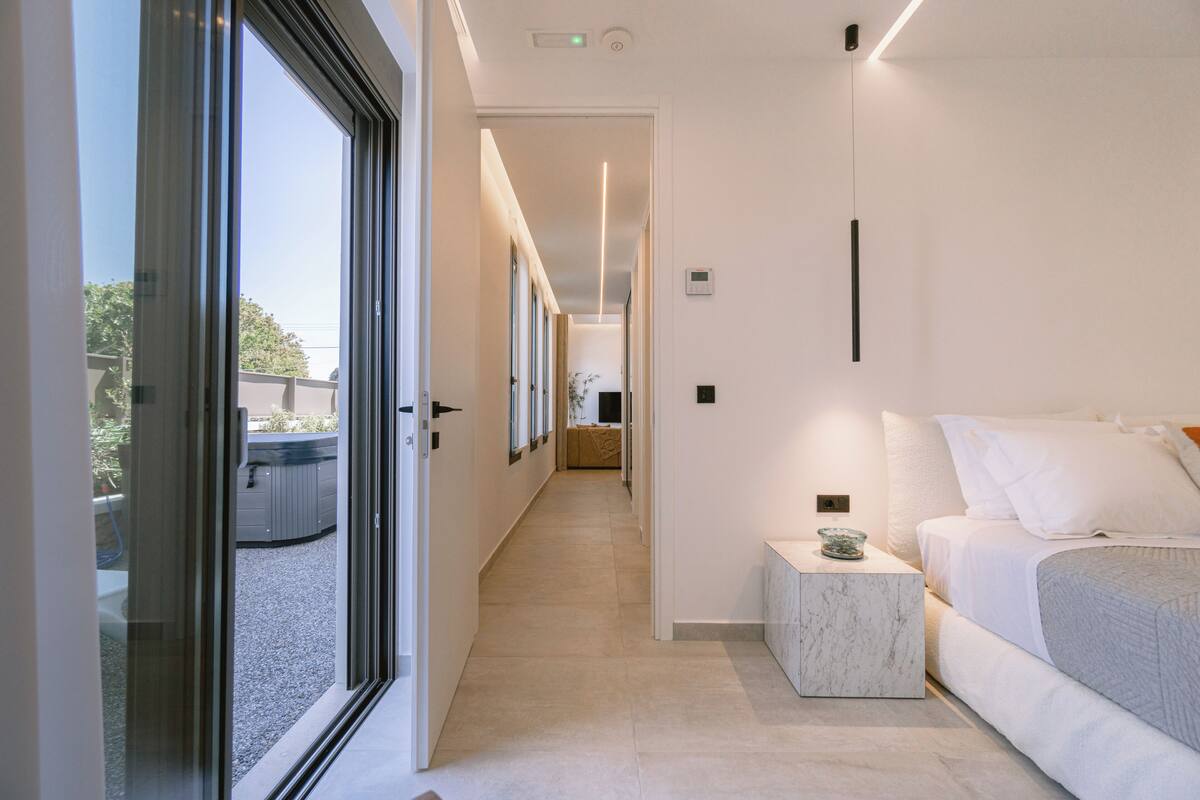 A modern hotel room with a large bed, a small bedside table, and a view through a glass door to an outdoor area.