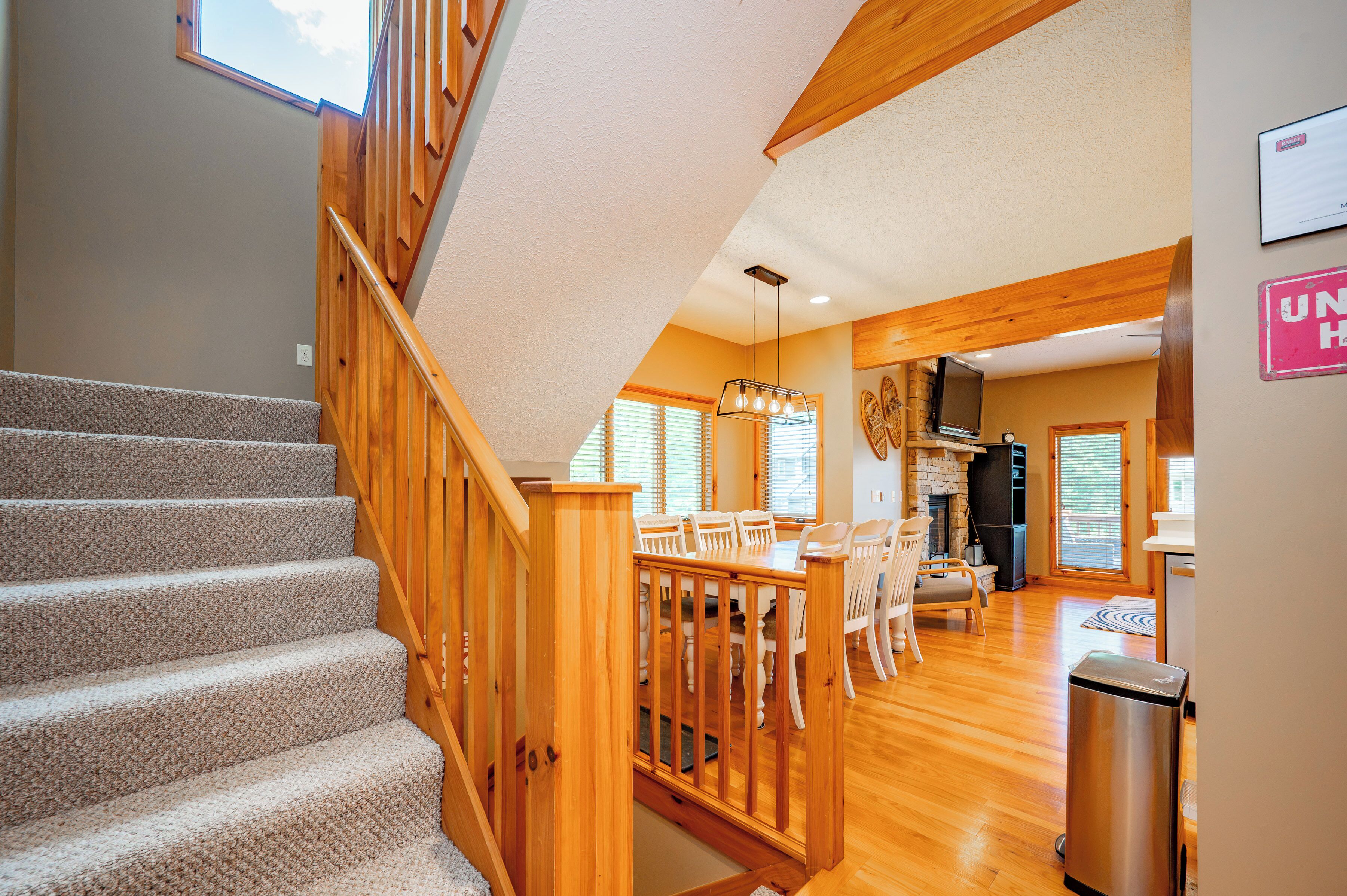 A staircase with wooden railings leading to a dining area with a fireplace and a kitchen area visible in the background.