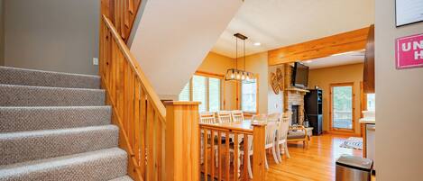 A staircase with wooden railings leading to a dining area with a fireplace and a kitchen area visible in the background.