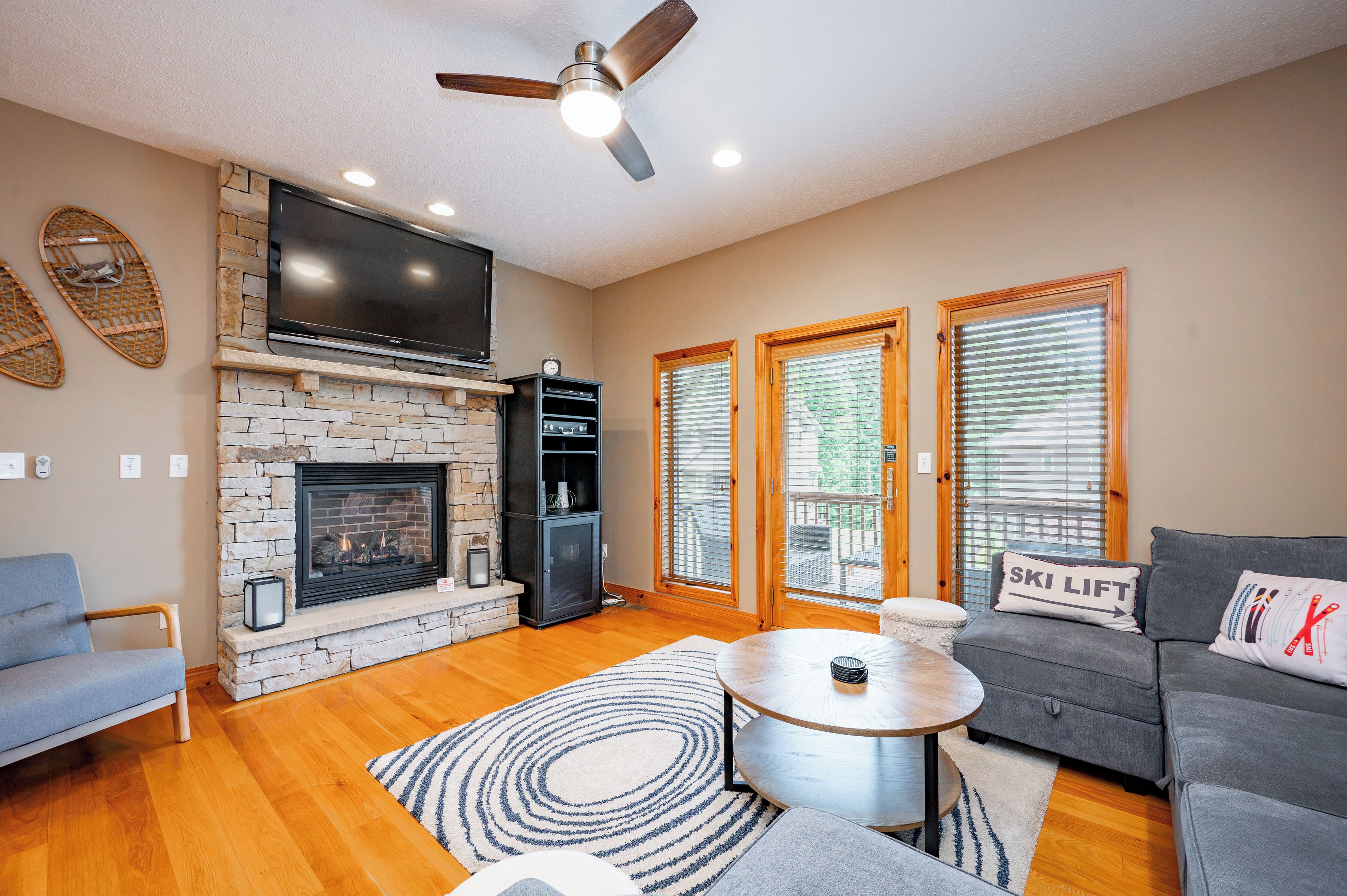 A living room with a fireplace, a flat-screen TV, a ceiling fan, and a sofa with a ski lift pillow.