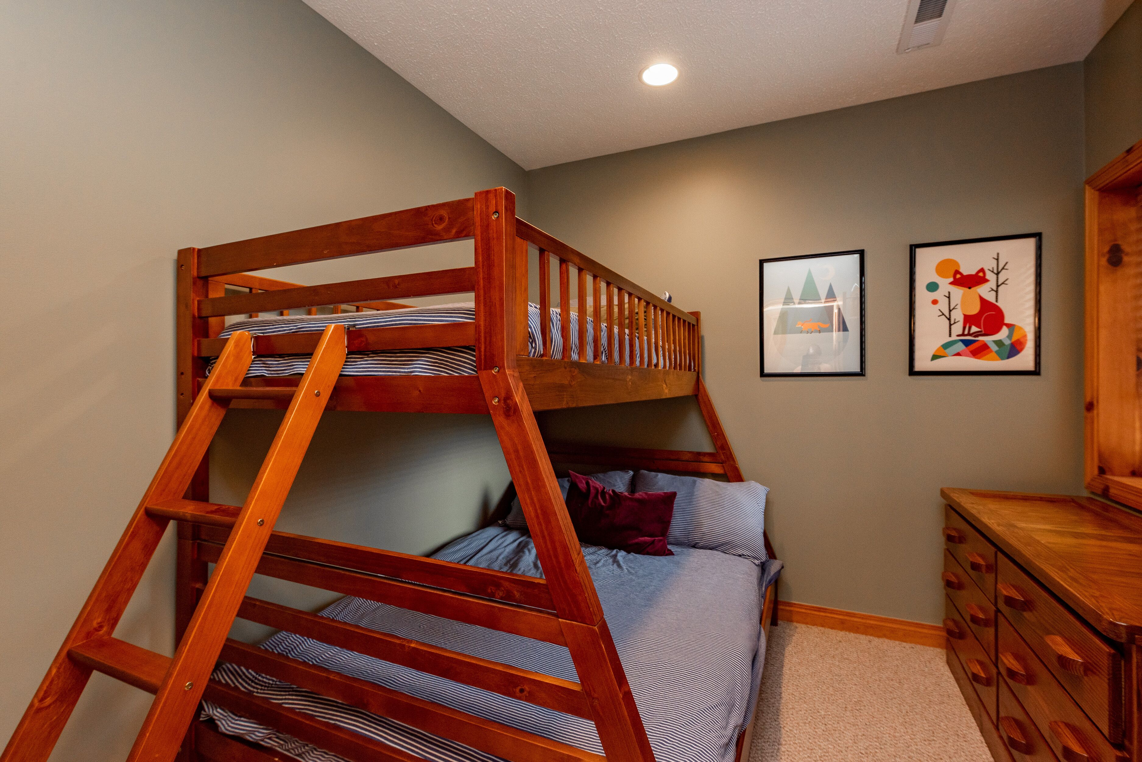 A bunk bed with a wooden ladder, a single bed with pillows, a dresser, and two framed pictures on the wall.