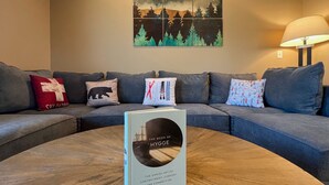A modern living room with a grey sofa, a round wooden coffee table, and a book titled "The Book of Hygge" on the table.