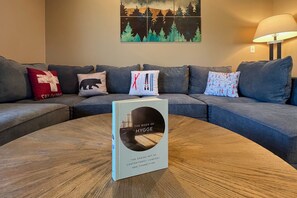 A modern living room with a grey sofa, a round wooden coffee table, and a book titled "The Book of Hygge" on the table. - Cozy Rustic Duplex w/ Hot Tub, Fireplace & Views (McHenry)