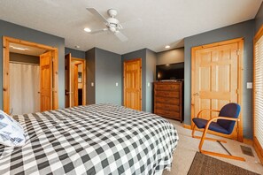 A bedroom with a checkered bedspread, a ceiling fan, wooden doors, a television, and a rocking chair. - Cozy Rustic Duplex w/ Hot Tub, Fireplace & Views (McHenry)