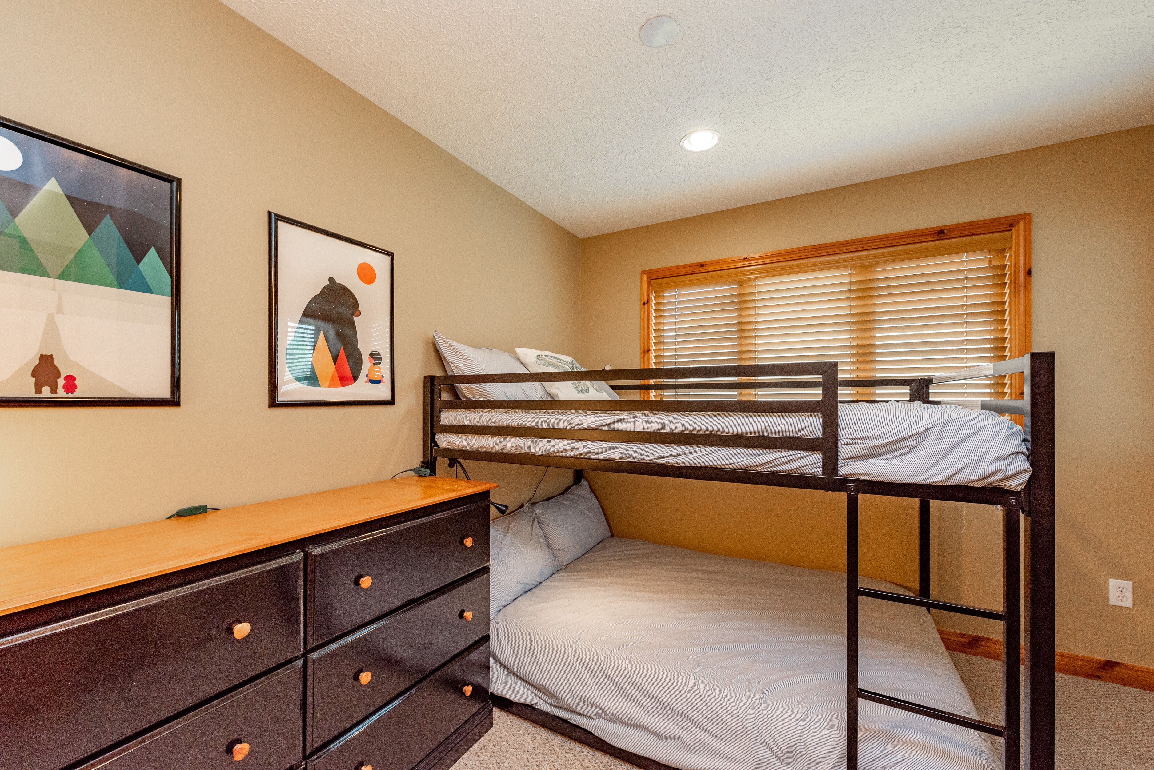 A bunk bed with a wooden headboard, a dresser with drawers, and a window with blinds.