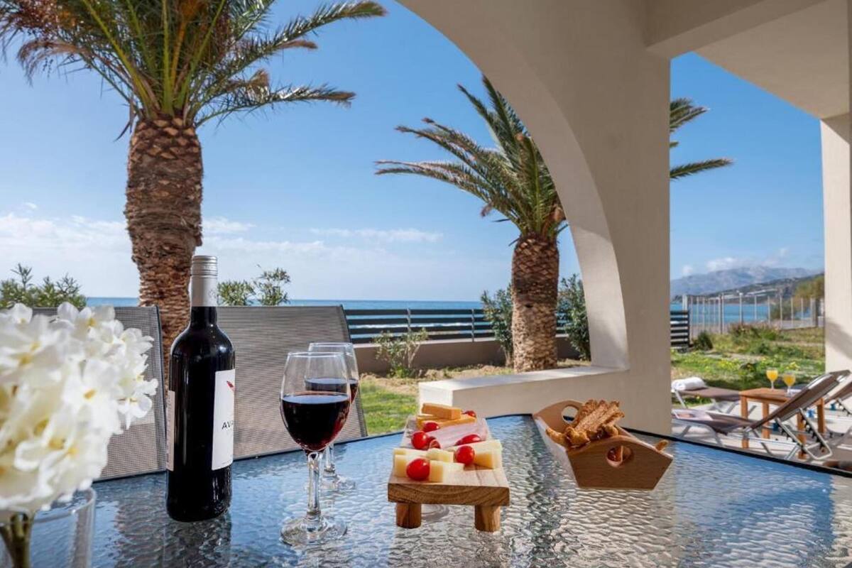 Uma mesa com garrafa de vinho, copos e tábua de queijos com frutas, com vista para uma praia com palmeiras e céu claro.