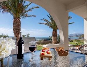 Uma mesa com garrafa de vinho, copos e tábua de queijos com frutas, com vista para uma praia com palmeiras e céu claro.