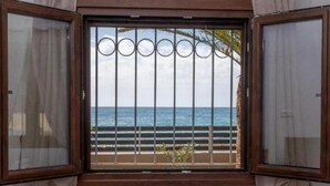 A bedroom with a large window overlooking the sea, a bed with an orange blanket, and a view of the beach and palm trees. - Atalanti Seaside Junior Suites (Viannos)