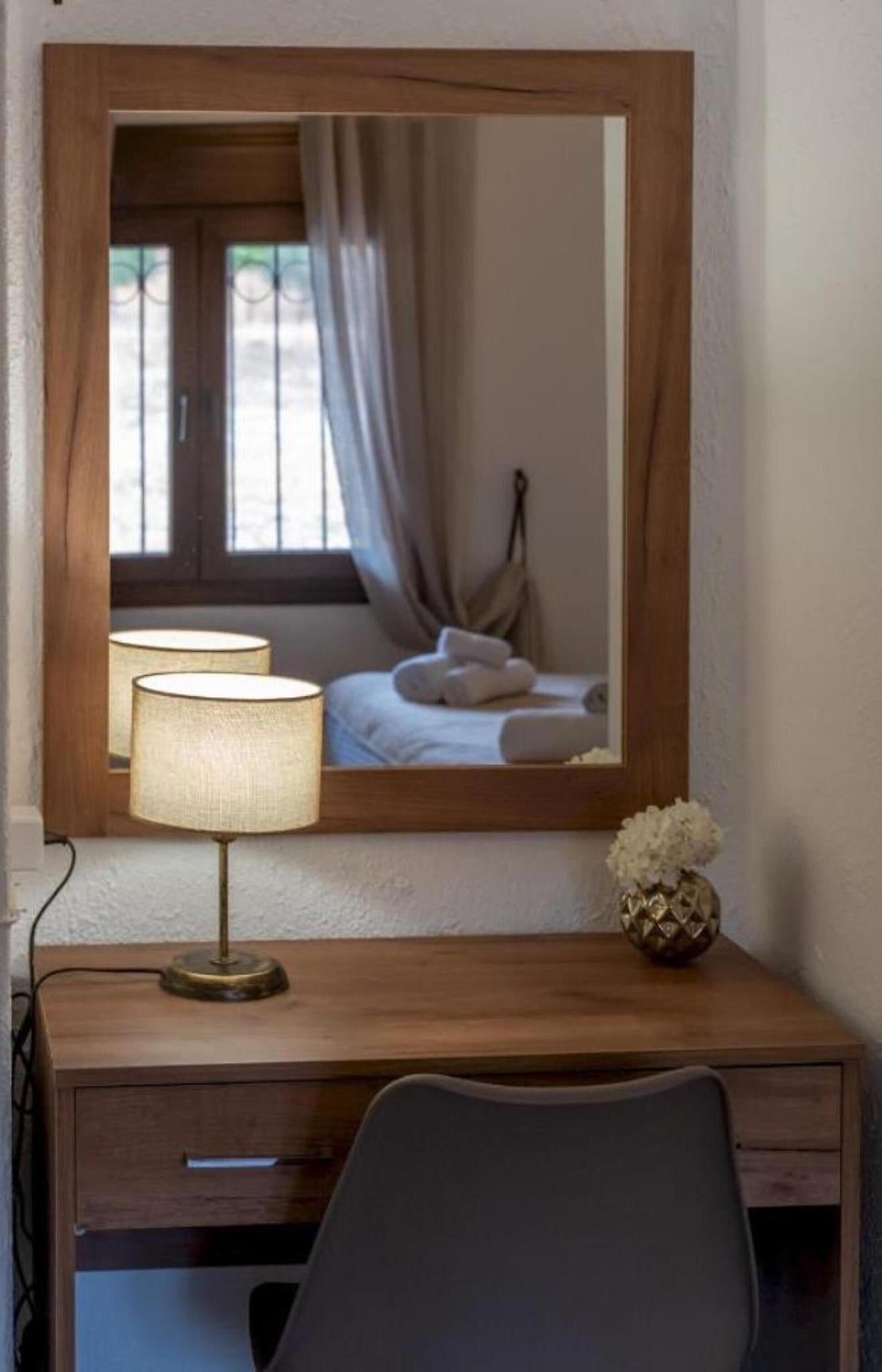 Una scrivania in legno con una lampada, uno specchio e un vaso con fiori bianchi.