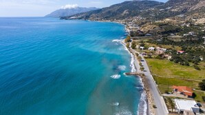 An aerial view of a coastal area with a clear blue sea, a road along the coast, and buildings in the background. - Atalanti Seaside Junior Suites (Viannos)