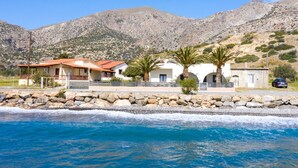 A coastal property with a stone wall, a building with a red roof, and a mountain in the background. - Atalanti Seaside Junior Suites (Viannos)