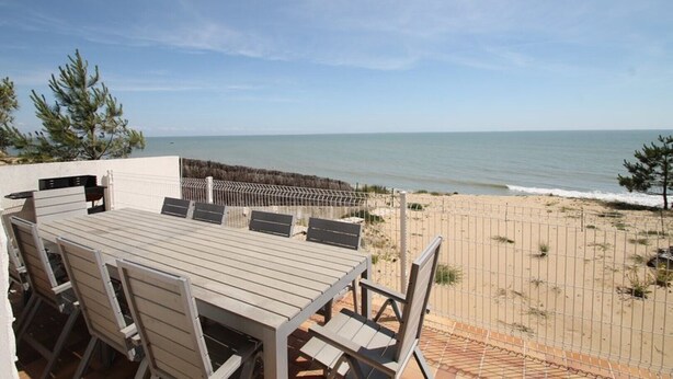 An outdoor patio with a wooden table and chairs, overlooking a beach with a fence separating the patio from the sand. - Beach House with 180 degree sea views, 6 Bed, 4 Baths, sleeps 10-12 (La Tranche Sur Mer)