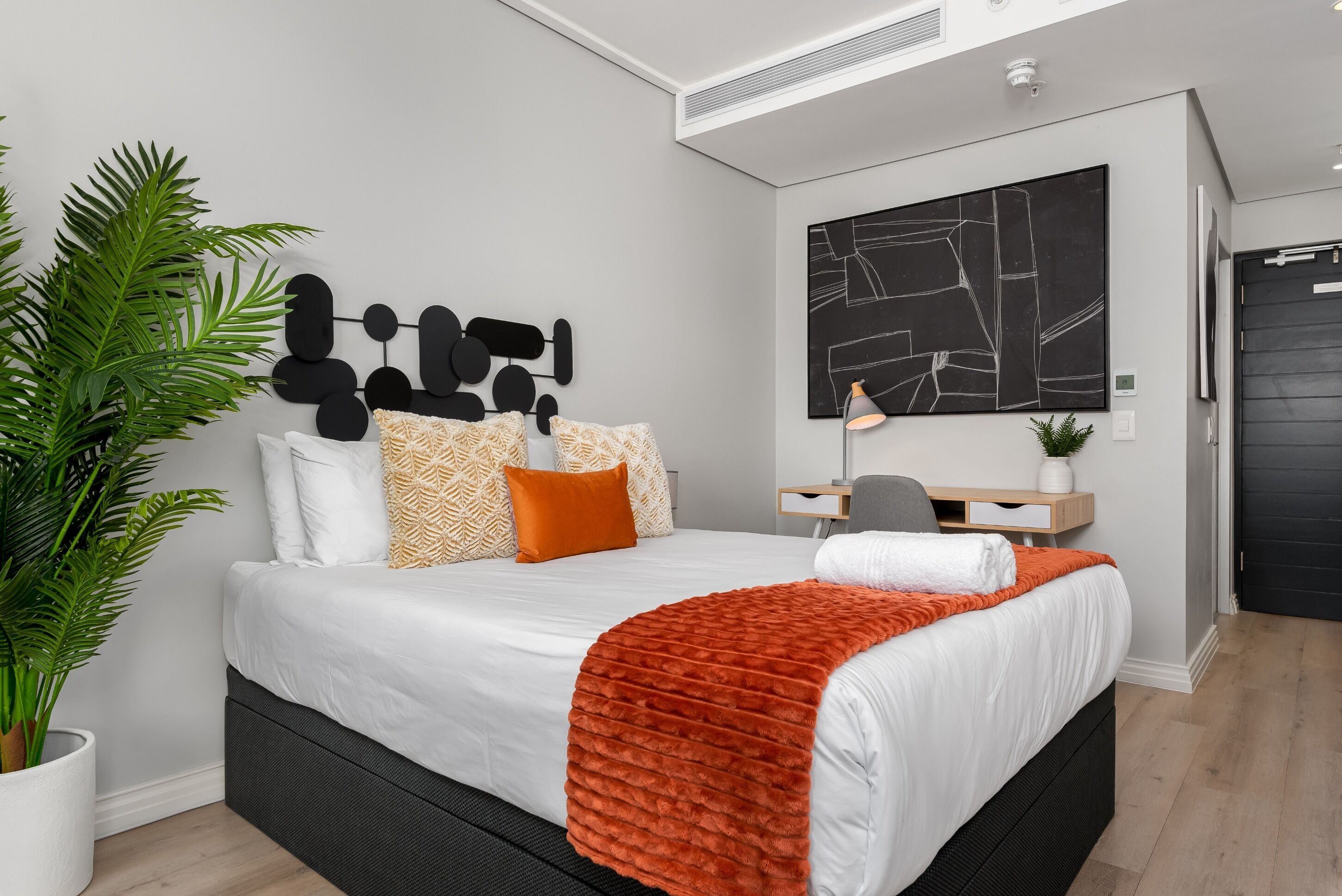 Un dormitorio moderno con una cama grande, una planta en maceta, un escritorio con una silla y un cuadro abstracto negro en la pared.