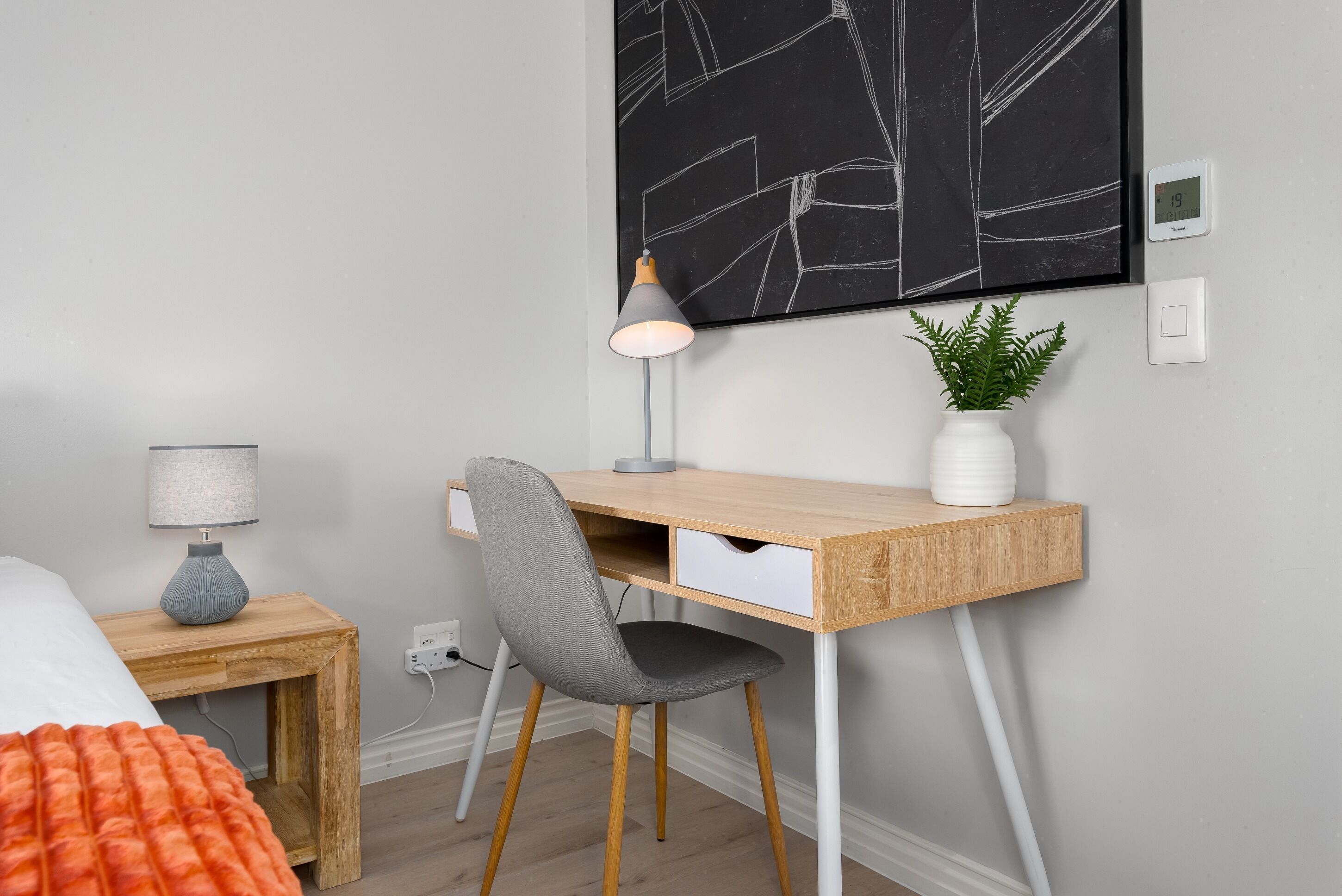 Un dormitorio moderno con un escritorio de madera, una silla gris, una mesita de noche con una lámpara y una planta en la pared.