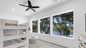 Un dormitorio moderno con literas, ventilador de techo, ventanas amplias con vistas a árboles y agua, y un otoman redondo.