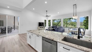 Una cocina moderna con una isla grande, muebles blancos y un lavavajillas. Hay una zona de comedor con una mesa y sillas, y vistas a un jardín a través de amplios ventanales.