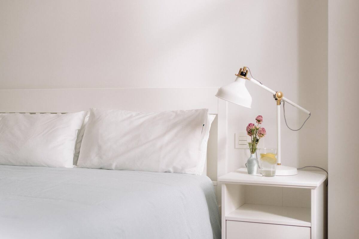 A neatly made bed with white bedding and pillows, a white bedside table with a lamp and a vase with flowers, and a wooden floor.