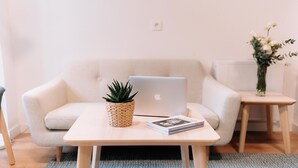A modern living room with a beige sofa, a small wooden table with a laptop and a book, a woven plant pot, and a vase with flowers. - San Bernardo 19 (Aviles)